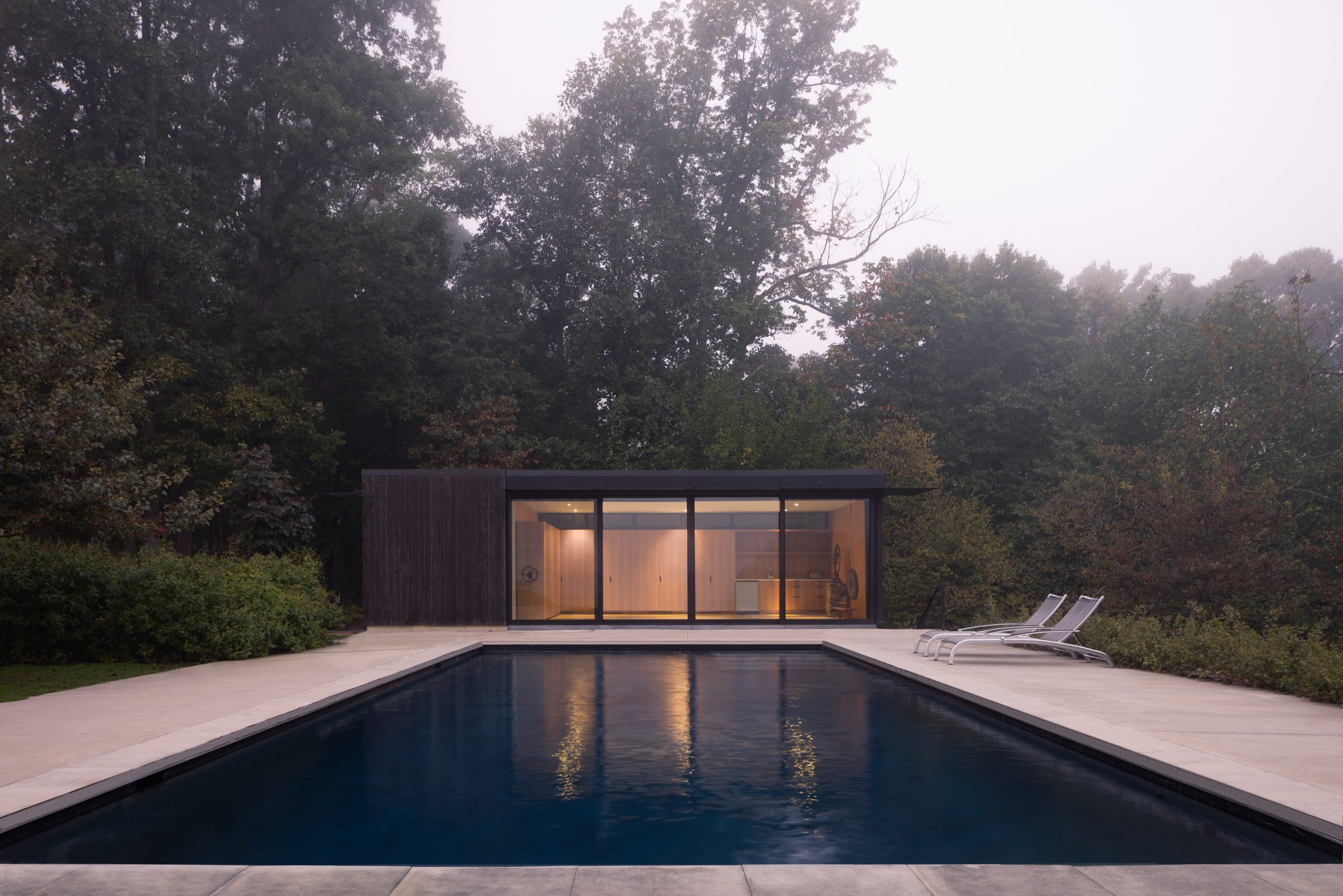 A glass-walled pool pavilion glowing at dusk in the fog, reflected in the still water