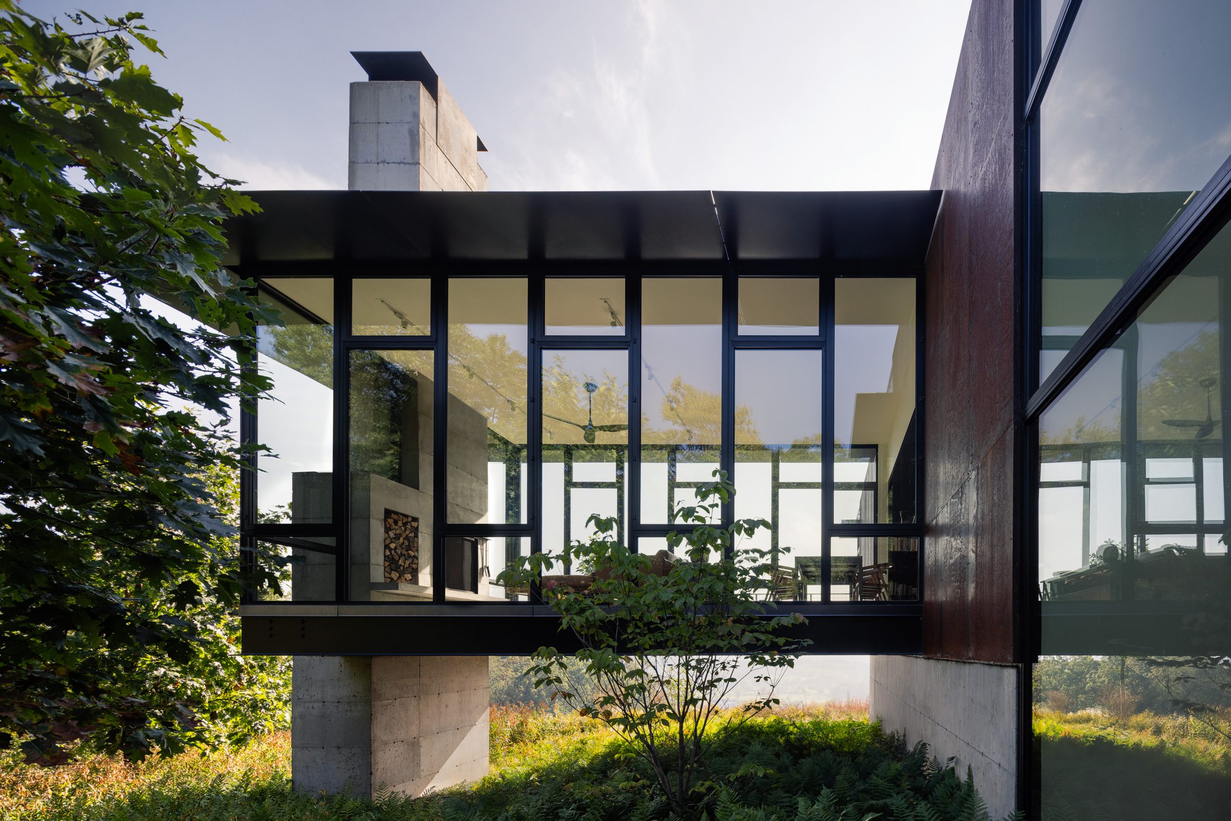 A modern glass-and-steel home open to the trees, warm interior light glowing through the walls