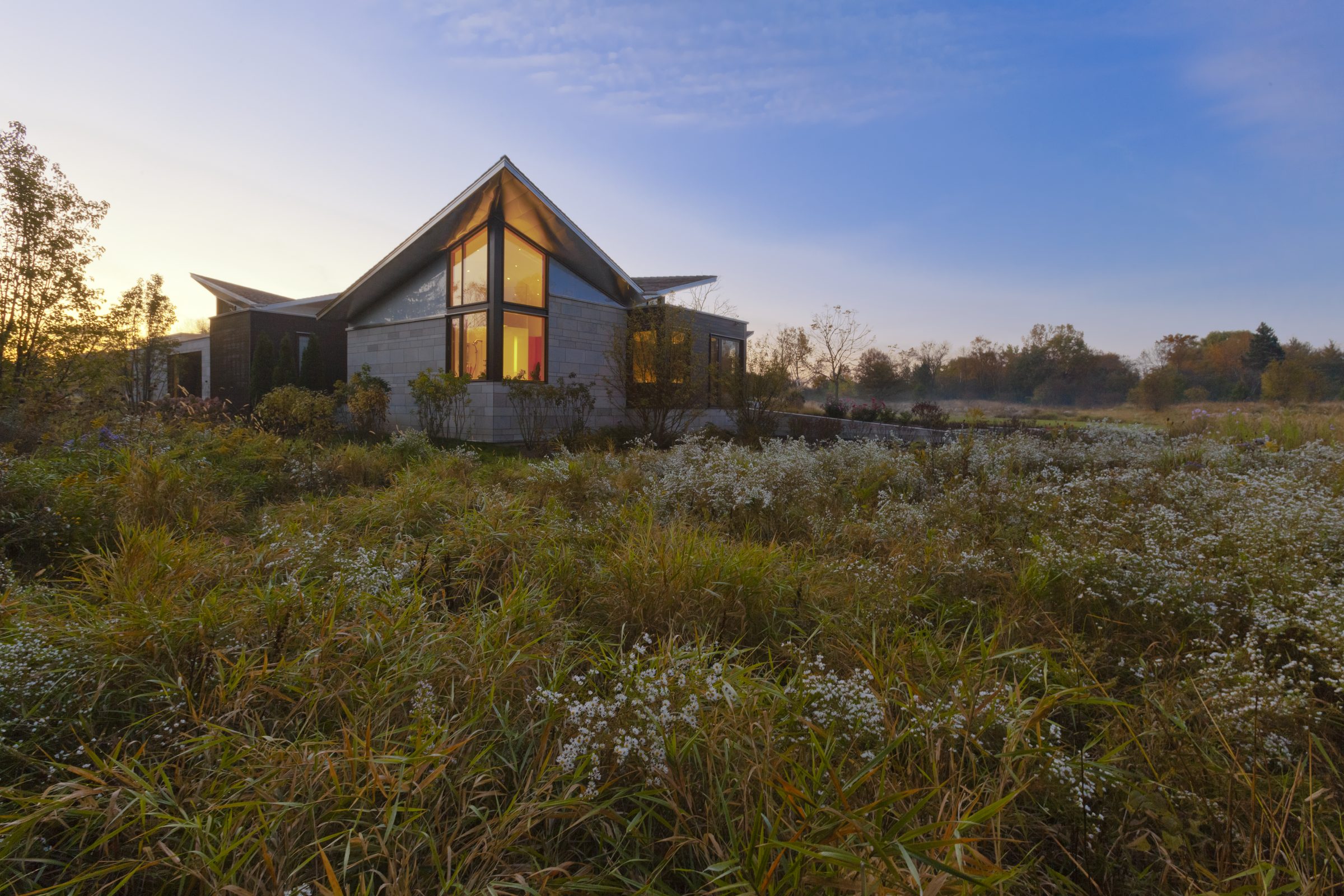 A modern glass-gabled home glowing warm at dusk in a wild meadow